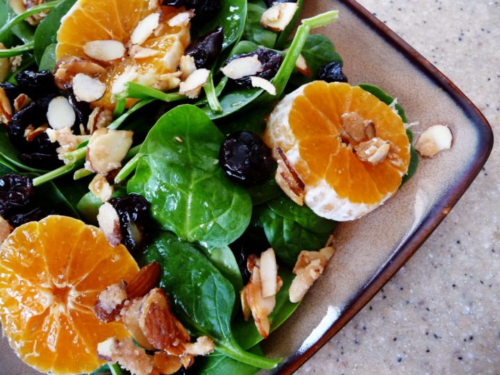 A vibrant spinach clementine salad on a rustic plate, featuring fresh baby spinach, juicy clementine slices, candied almonds, and dried cherries, all lightly dressed for a refreshing and flavorful dish