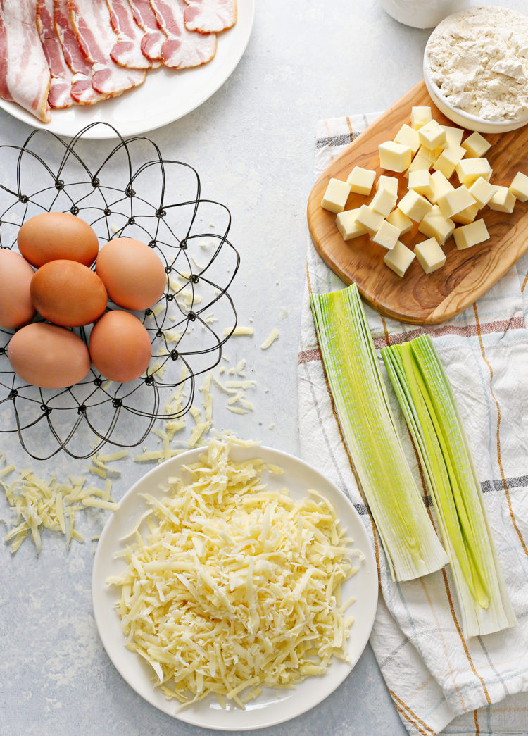 cleaned leeks on a dish towel next to a basket of eggs, a plate of bacon, and a plate of cheese