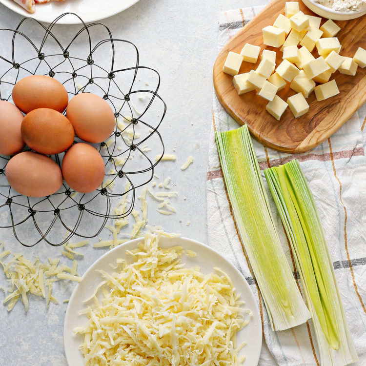 cleaned leeks on a dish towel next to a basket of eggs, a plate of bacon, and a plate of cheese