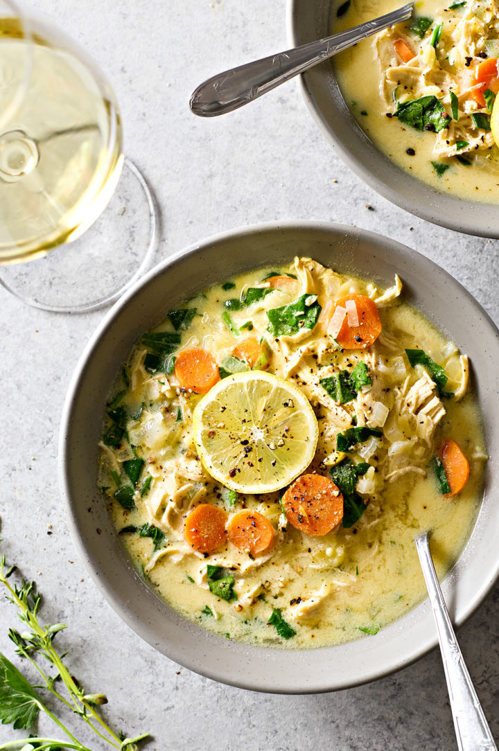 Overhead view of two bowls of creamy chicken spinach orzo soup, each with a spoon, garnished with lemon slices, and accompanied by a glass of white wine, fresh thyme, and parsley on a grey background