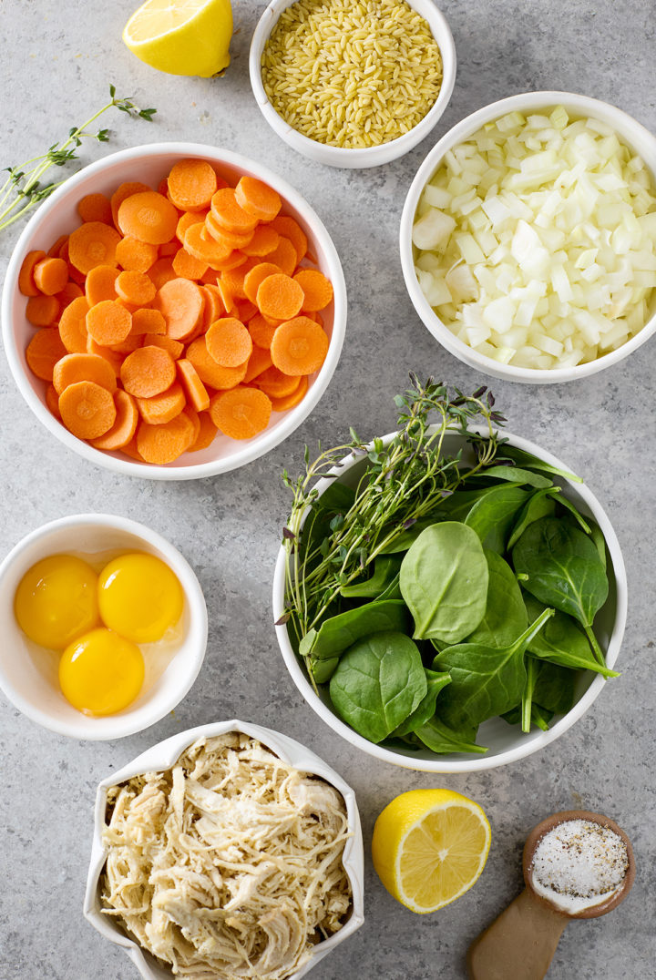 Ingredients for chicken spinach orzo soup: orzo pasta, chicken, spinach, carrots, onion, egg yolk, lemon, and fresh herbs on a countertop.