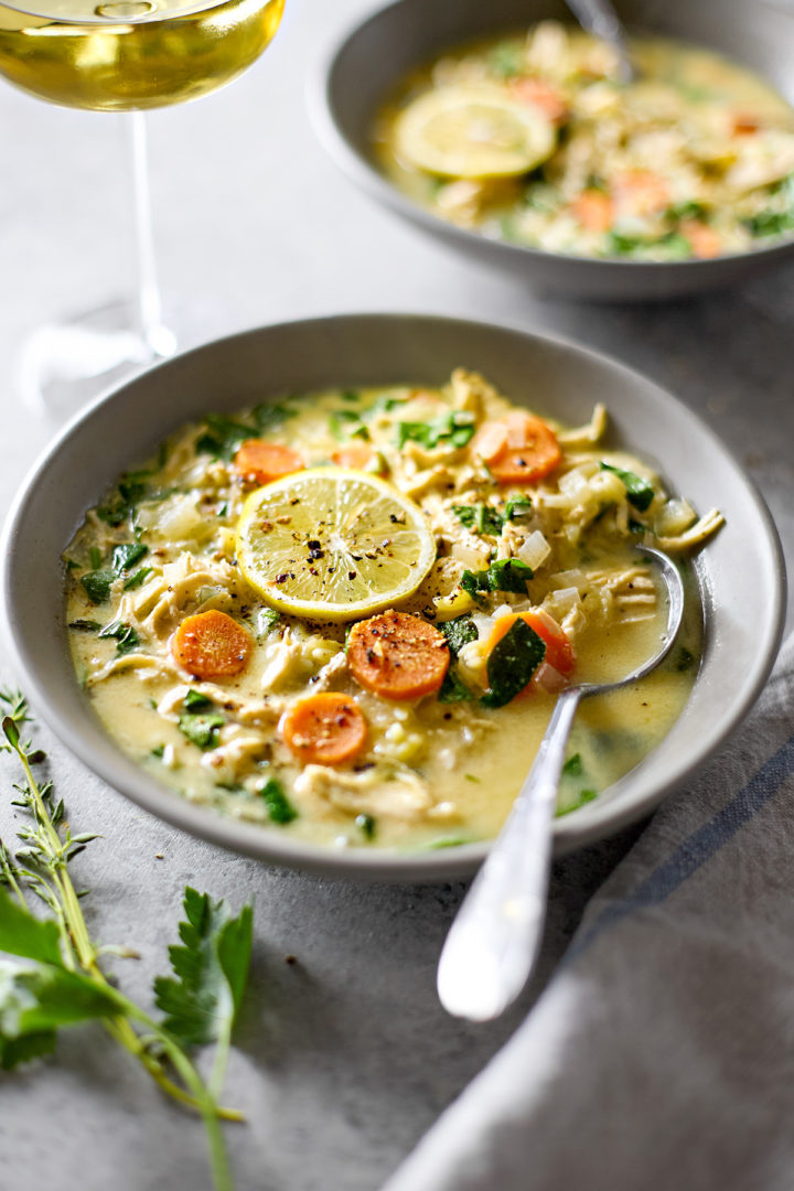 Two bowls of creamy chicken spinach orzo soup garnished with lemon slices, served with a glass of white wine, fresh thyme, and parsley on a grey background