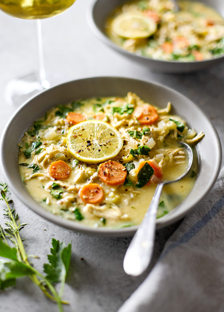 Two warm bowls of bowl of lemon chicken spinach orzo soup with fresh lemon, parsley, and thyme garnishes served with a glass of white wine.