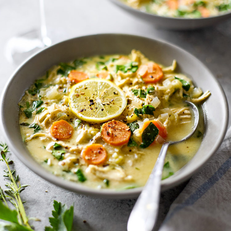 Two warm bowls of bowl of lemon chicken spinach orzo soup with fresh lemon, parsley, and thyme garnishes served with a glass of white wine.