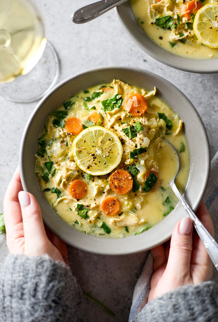 Overhead flat lay of a woman's hands holding a bowl of creamy chicken spinach orzo soup, wearing a cozy grey sweater