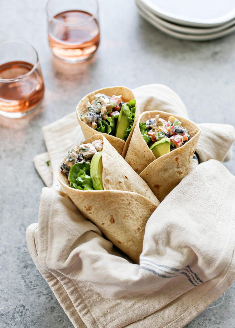 A linen-lined basket holds three Southwest Chicken Salad Wraps with creamy chicken salad, lettuce, and avocado. Two glasses of rosé and white plates sit in the background on a gray surface.