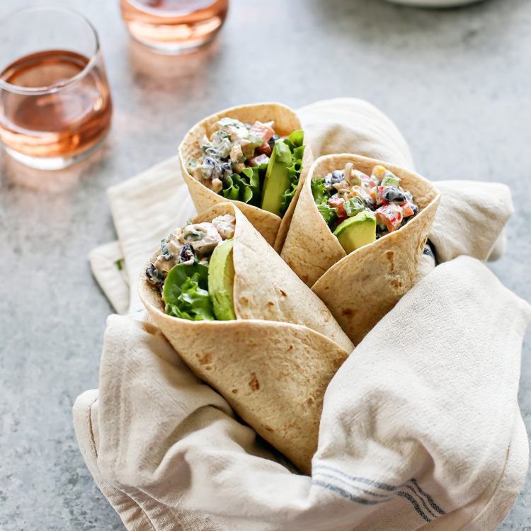 A linen-lined basket holds three Southwest Chicken Salad Wraps with creamy chicken salad, lettuce, and avocado. Two glasses of rosé and white plates sit in the background on a gray surface.