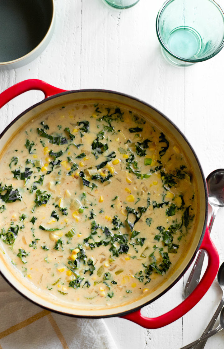 A red Dutch oven filled with creamy roasted corn chowder, featuring chunks of potato, kale, and golden roasted corn in a velvety broth. The soup is ready to be served with ladles and bowls nearby.