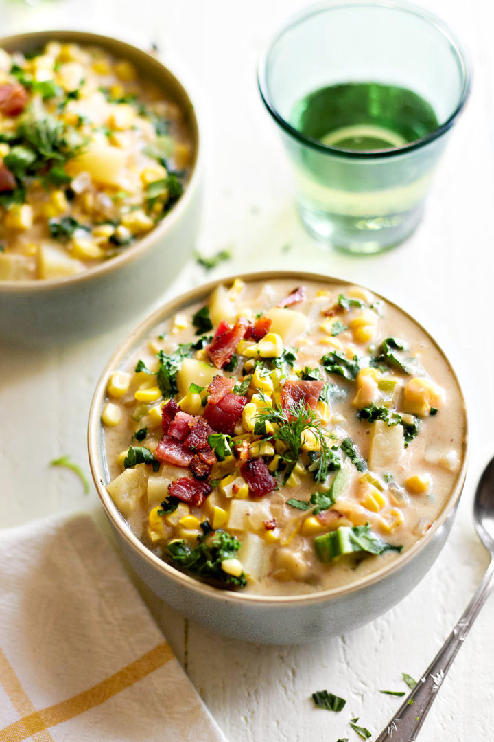 A bowl of roasted corn chowder with potatoes and bacon, topped with crispy bacon bits and fresh herbs. The creamy soup is filled with golden roasted corn, tender potatoes, and kale, served in a ceramic bowl on a white table.