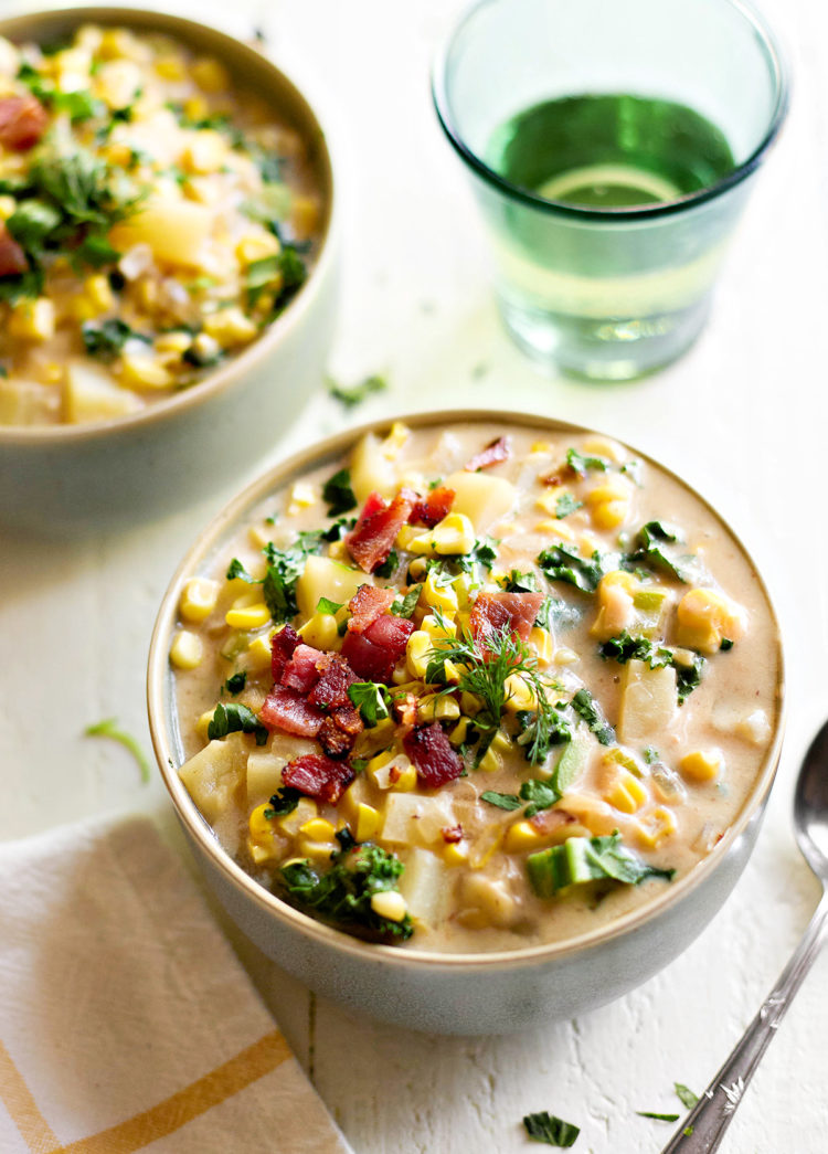 A close-up of bacon potato corn chowder served in a bowl on a white wooden table. The creamy broth is filled with tender potatoes, sweet roasted corn, and crispy bacon, garnished with fresh parsley and dill. A spoon rests beside the bowl, ready for serving.