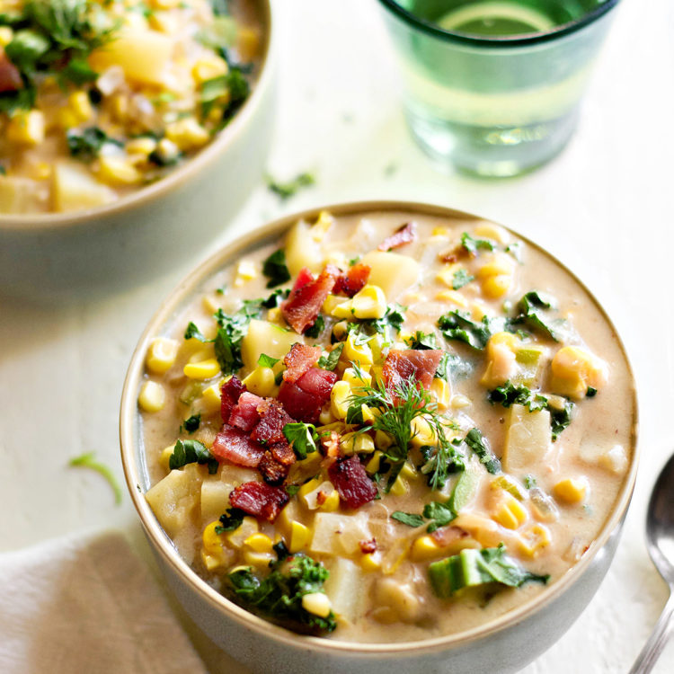 A close-up of bacon potato corn chowder served in a bowl on a white wooden table. The creamy broth is filled with tender potatoes, sweet roasted corn, and crispy bacon, garnished with fresh parsley and dill. A spoon rests beside the bowl, ready for serving.