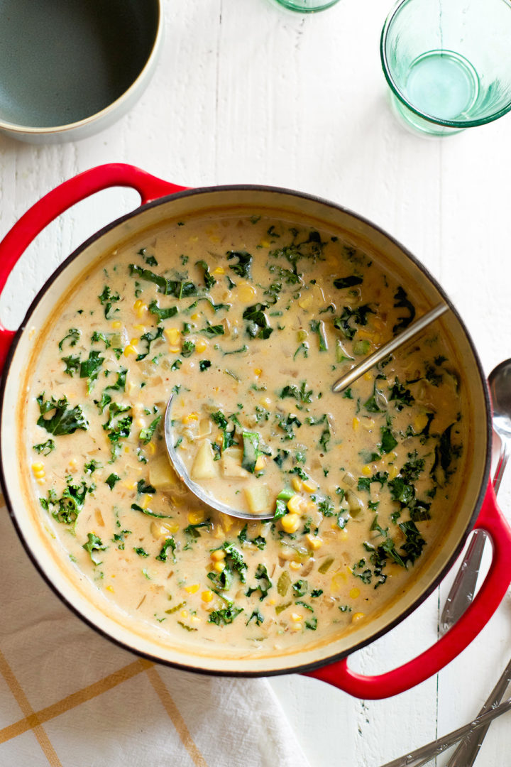 A red Dutch oven filled with corn and potato chowder with bacon, featuring chunks of potato, kale, and golden roasted corn in a velvety broth. A ladle rests inside the pot, ready for serving.