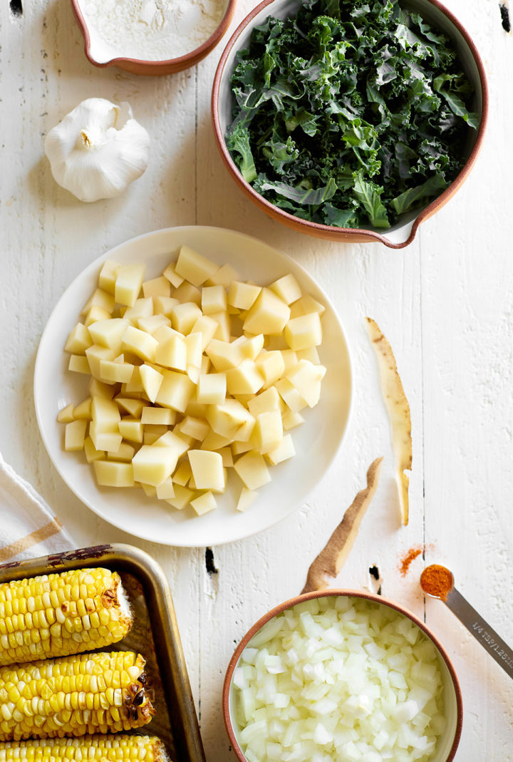 A flat lay of fresh ingredients for roasted corn chowder, including diced potatoes, chopped kale, roasted corn on the cob, diced onions, garlic, and a small dish of cayenne pepper, arranged on a white wooden surface.