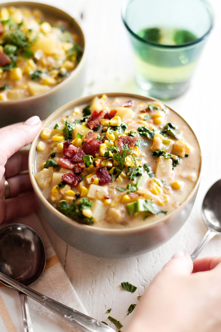 A woman’s hands holding a bowl of roasted corn and potato chowder, ready to serve. The soup is rich and creamy, with pieces of crispy bacon, roasted corn, and tender potatoes visible on top.