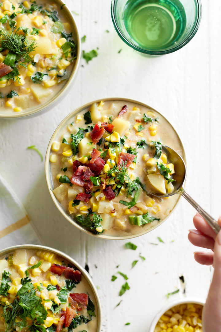 A spoonful of creamy roasted corn chowder being lifted from a bowl, showing chunks of golden corn, potatoes, and crispy bacon in a rich, flavorful broth.