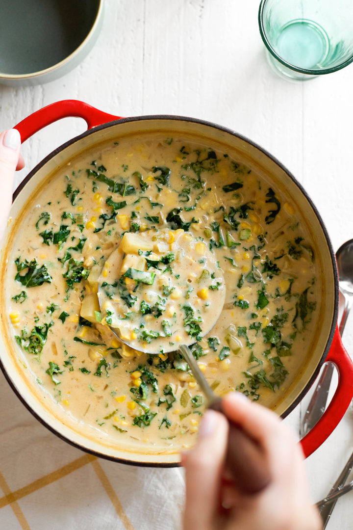 A woman holds a red Dutch oven filled with creamy roasted corn and potato chowder, while using a ladle to serve the soup. The rich broth is packed with golden roasted corn, tender potatoes, and crispy bacon, creating a warm and hearty meal.