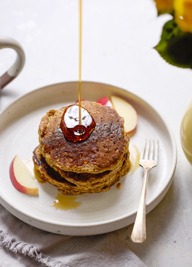 A stack of golden-brown oatmeal applesauce pancakes on a plate, topped with syrup being drizzled, with apple slices on the side.