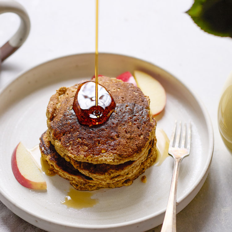 A stack of golden-brown oatmeal applesauce pancakes on a plate, topped with syrup being drizzled, with apple slices on the side.