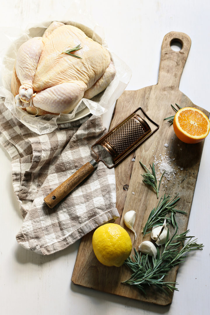 Ingredients for making roasted chicken for quinoa chicken salad, including a whole raw chicken on a plate alongside a wooden cutting board with fresh rosemary, lemon, orange, garlic cloves, and salt."