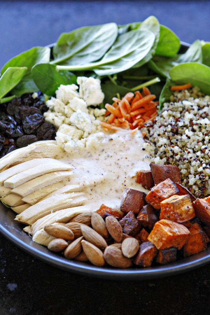 A vibrant quinoa chicken salad served in a bowl, featuring fresh baby spinach, roasted sweet potatoes, shredded chicken, cooked quinoa, blue cheese crumbles, dried cherries, and whole almonds, all drizzled with a creamy almond white balsamic dressing.