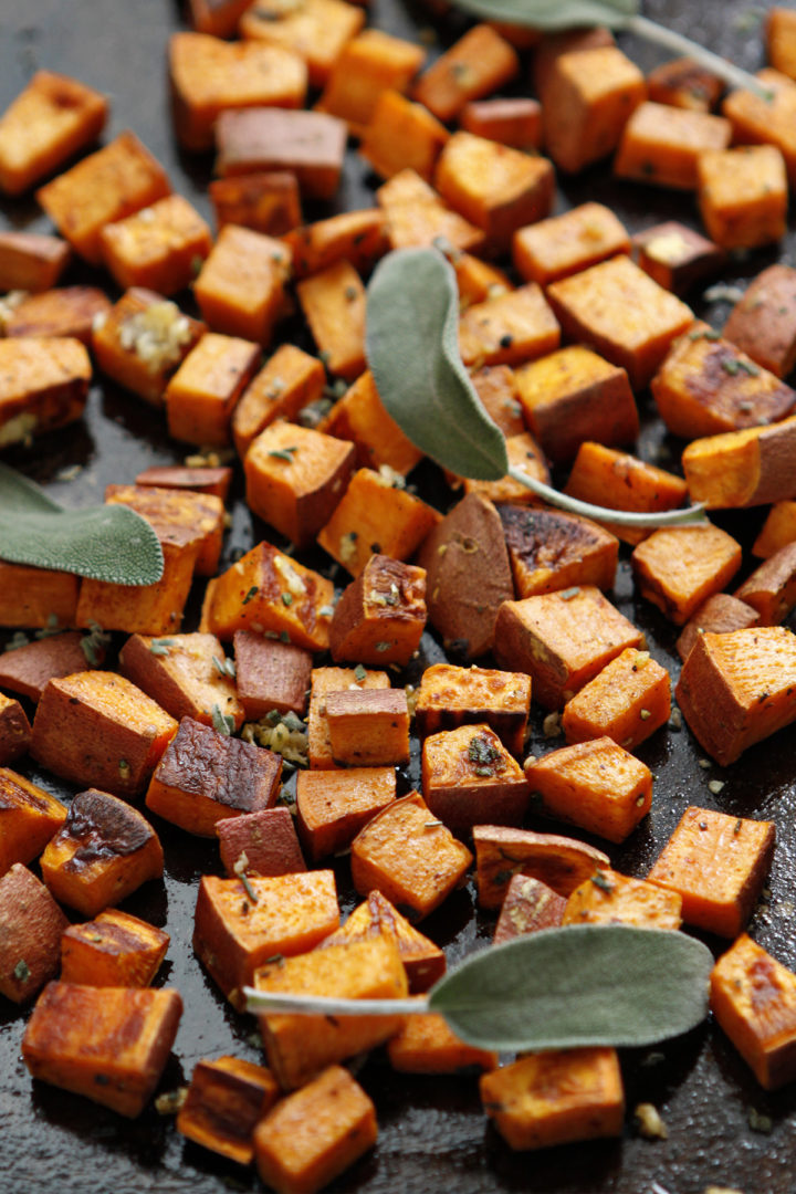 Perfectly roasted sweet potatoes, golden brown and tender, tossed with minced fresh garlic and chopped sage on a baking sheet—ready to be added to the spinach quinoa chicken salad for extra flavor.