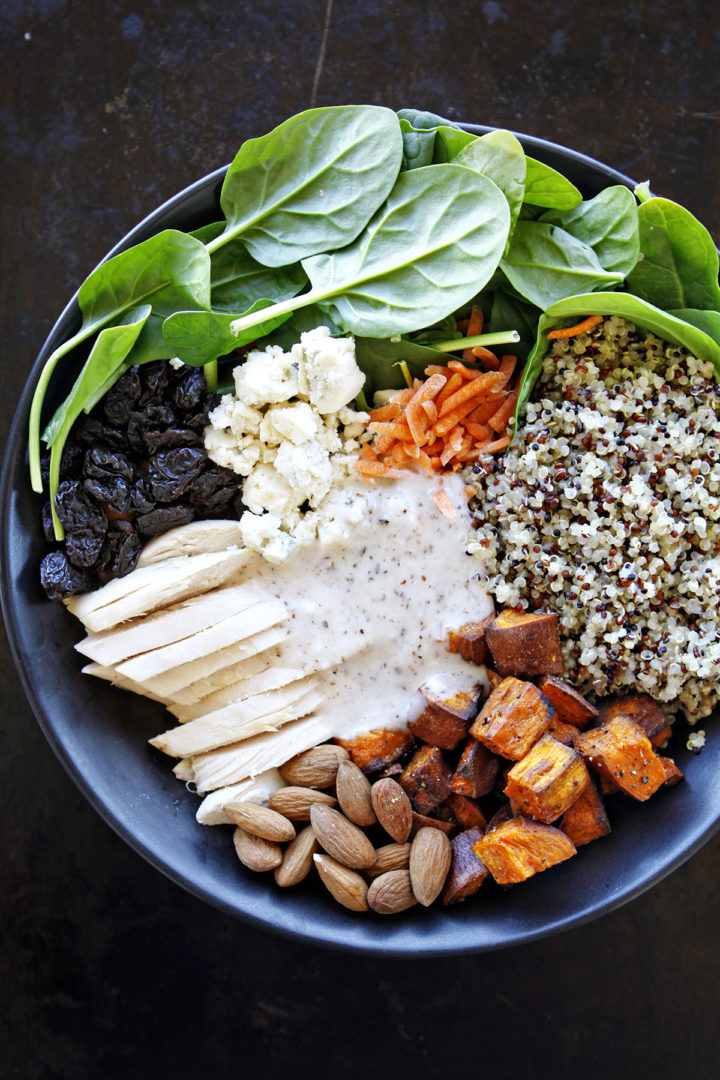 An overhead view of a spinach and quinoa salad with chicken, featuring a colorful mix of fresh baby spinach, roasted sweet potatoes, shredded chicken, fluffy quinoa, blue cheese crumbles, dried cherries, and crunchy almonds, topped with a creamy almond white balsamic dressing.