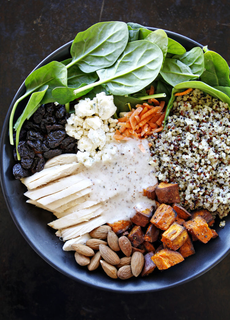 A vibrant quinoa chicken salad served in a bowl, featuring fresh baby spinach, roasted sweet potatoes, shredded chicken, cooked quinoa, blue cheese crumbles, dried cherries, and whole almonds, all drizzled with a creamy almond white balsamic dressing