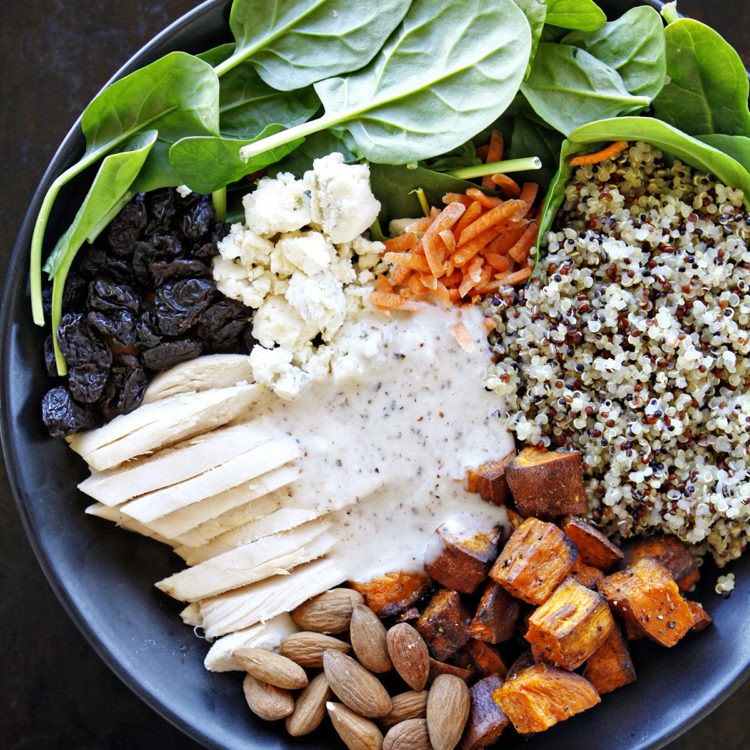 A vibrant quinoa chicken salad served in a bowl, featuring fresh baby spinach, roasted sweet potatoes, shredded chicken, cooked quinoa, blue cheese crumbles, dried cherries, and whole almonds, all drizzled with a creamy almond white balsamic dressing