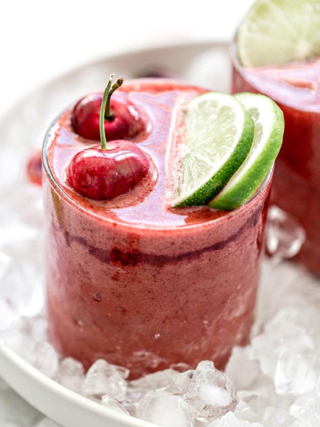 close up cherry slushie in a clear glass with fresh cherries and limes