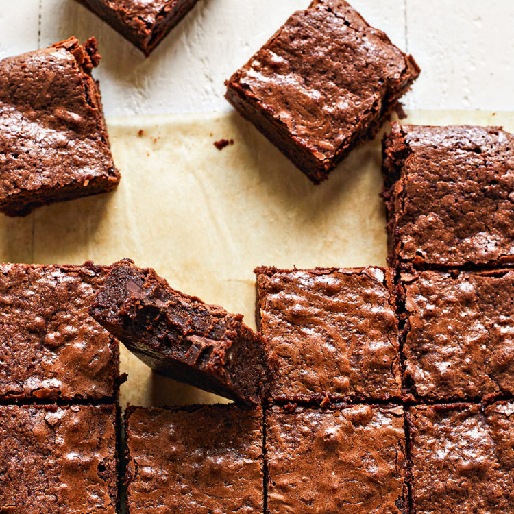 Overhead shot of cut squares of fudgy homemade brownies arranged on brown parchment paper on a white wooden table. One brownie with a bite taken out of it is placed on its side among the other brownies