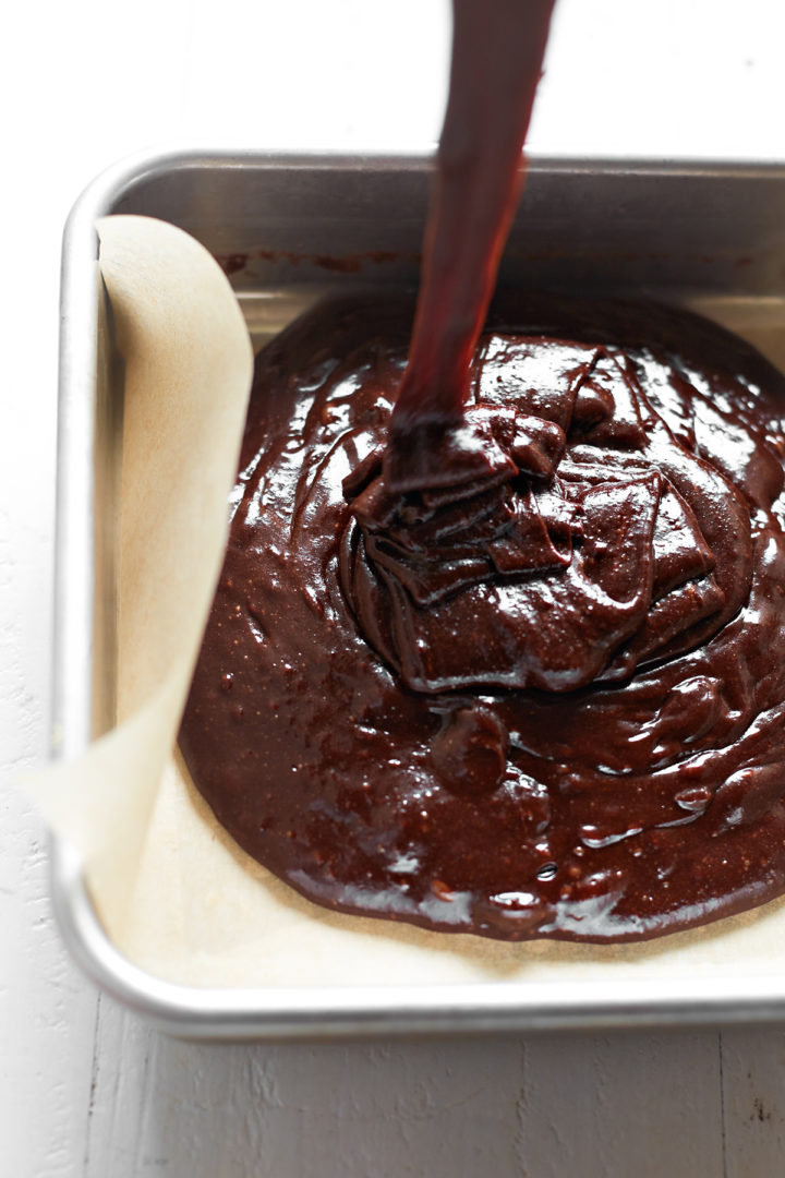 Thick, fudgy brownie batter being poured into a parchment-lined metal baking pan on a white wooden background.