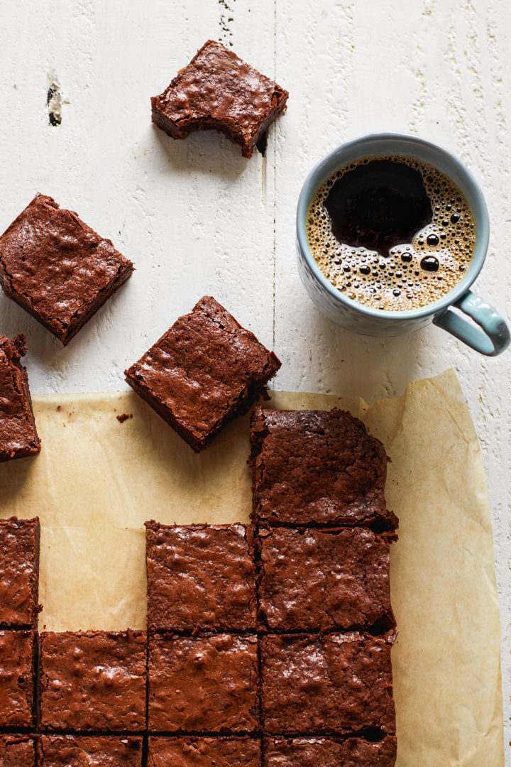 Overhead shot of cut squares of fudgy homemade brownies arranged on brown parchment paper on a white wooden table. One brownie has a bite taken out of it and sits next to a mug of hot coffee.