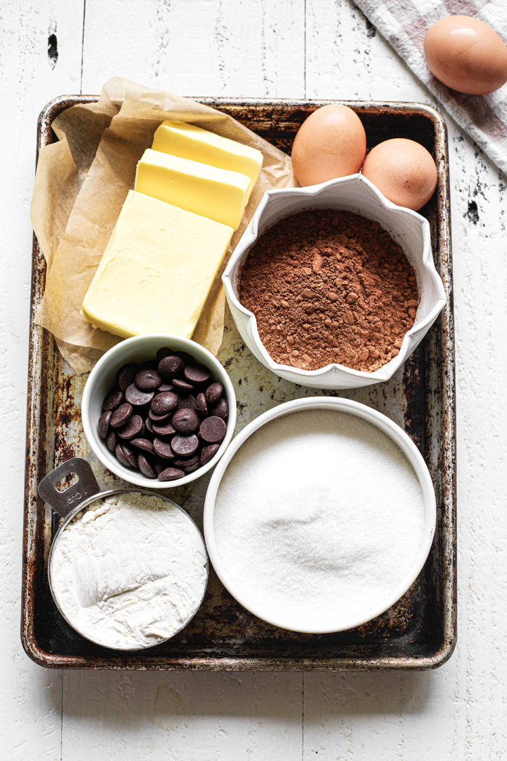 A baking sheet with bowls of ingredients for making homemade brownies, including butter, cocoa powder, sugar, and flour.