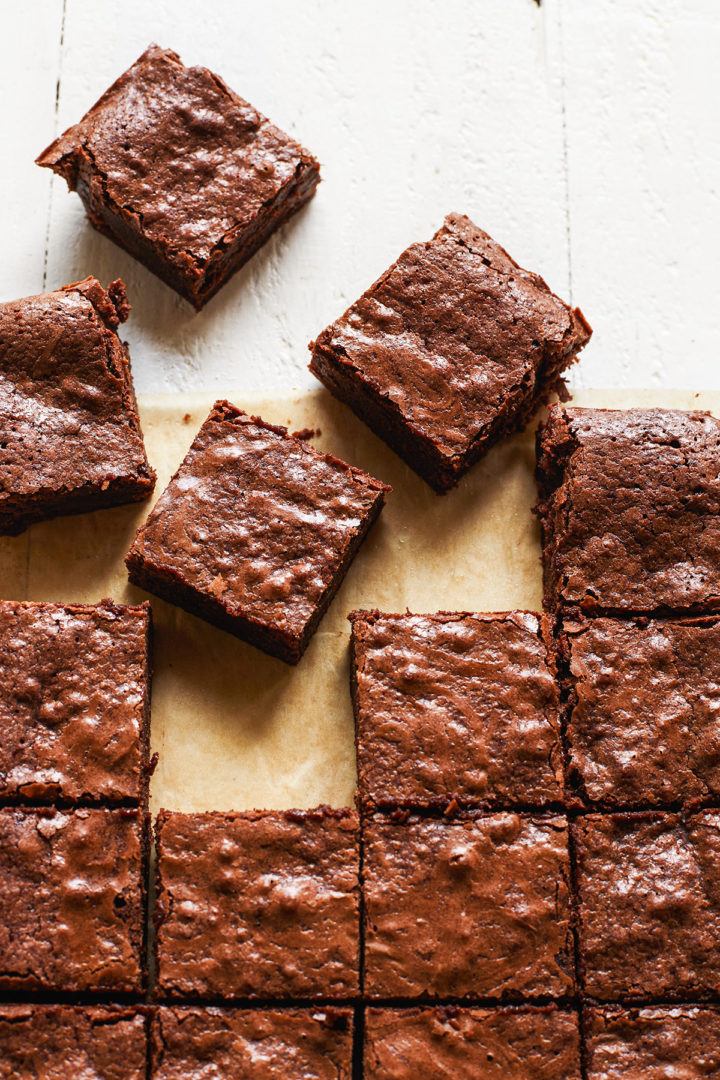 Overhead shot of cut squares of fudgy homemade brownies artfully arranged on brown parchment paper on a white wooden table. 