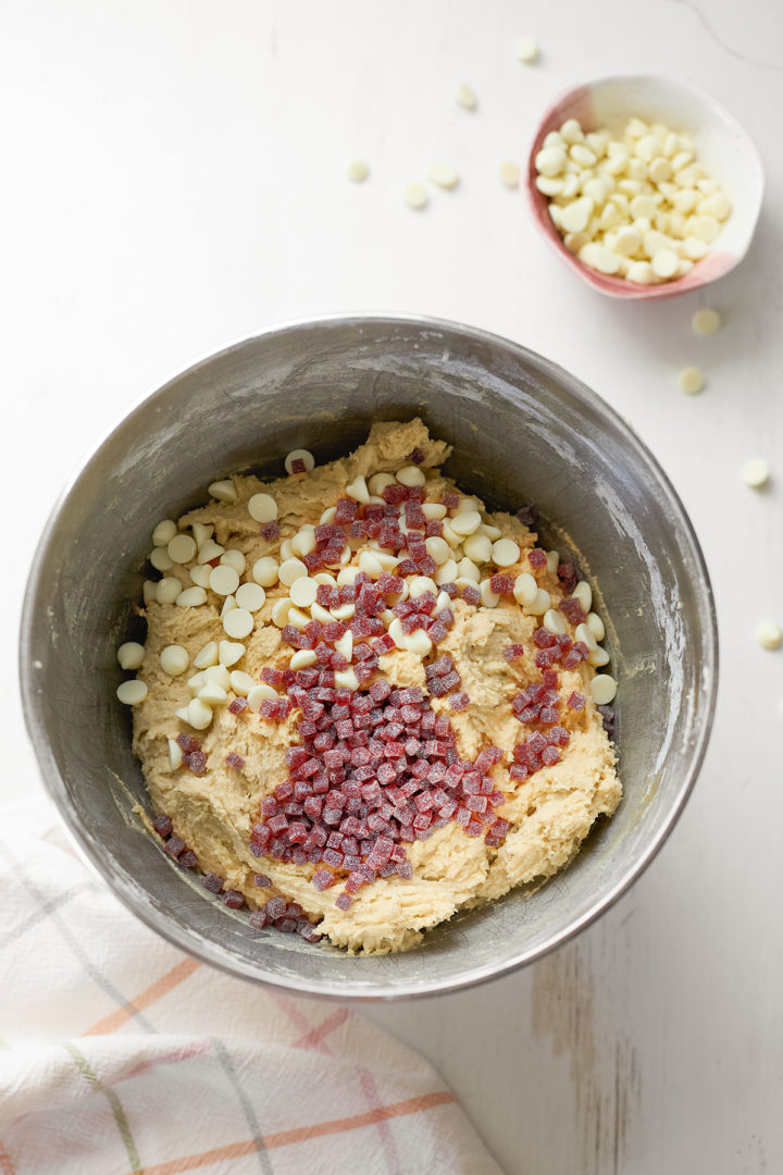 A mixing bowl filled with cookie dough, topped with white chocolate chips and raspberry jammy bits, ready to be mixed. A small bowl of extra white chocolate chips sits nearby on a white surface.