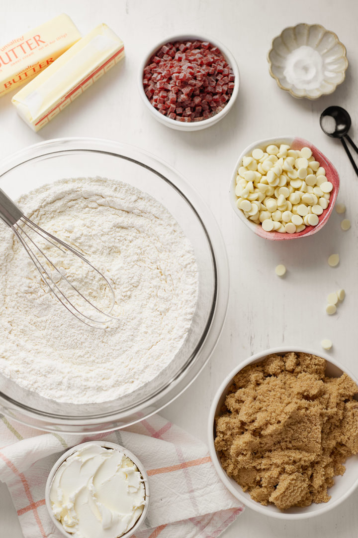 A flat lay of ingredients for Subway raspberry cheesecake cookies, including flour, brown sugar, cream cheese, butter, white chocolate chips, and raspberry jammy bits, arranged on a white countertop.