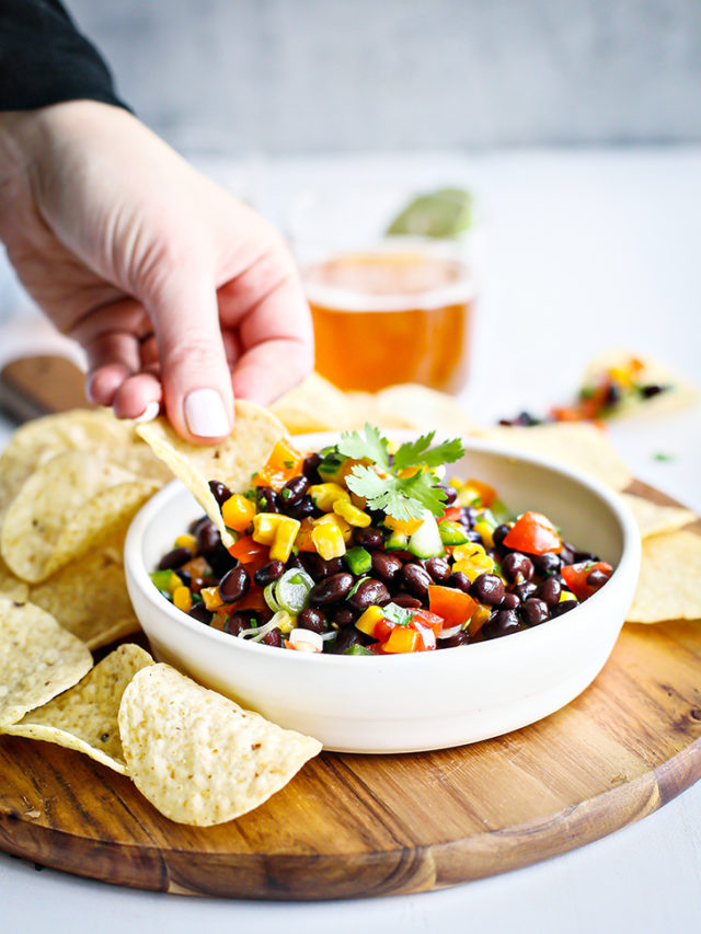 photo of black bean salad in a white bowl with woman scooping with a tortilla chip