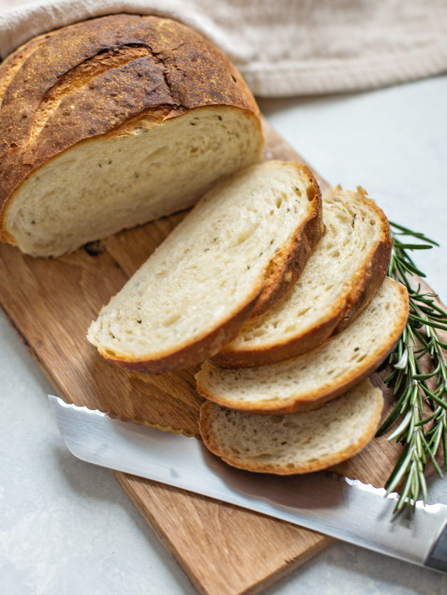 rosemary garlic bread on a wooden cutting board with a bread knife and sprig of fresh rosemary