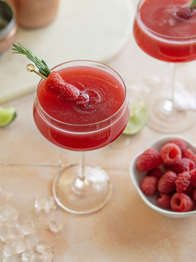 two glasses of a raspberry cosmopolitan next to a bowl of raspberries and slices of lime