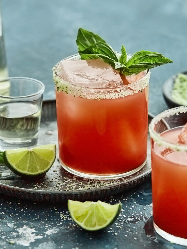 3 glasses of strawberry basil margarita on a teal background with a bottle of tequila, fresh lime wedges and basil sprigs