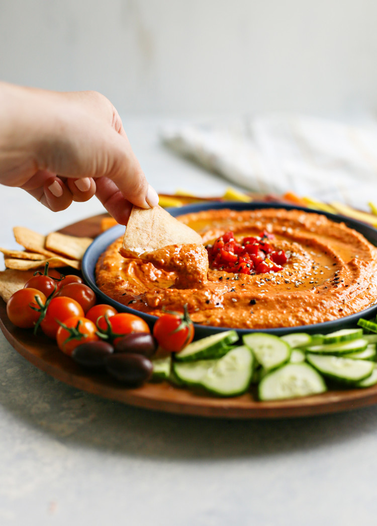 woman scooping red pepper hummus with a pita chip