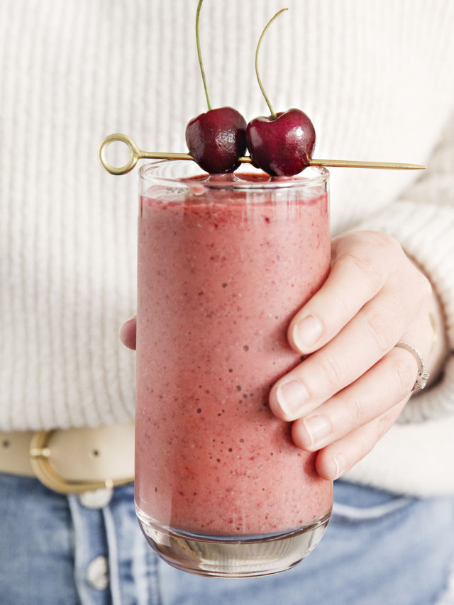 woman holding a glass of mango cherry smoothie garnished with fresh cherries on a cocktail skewer