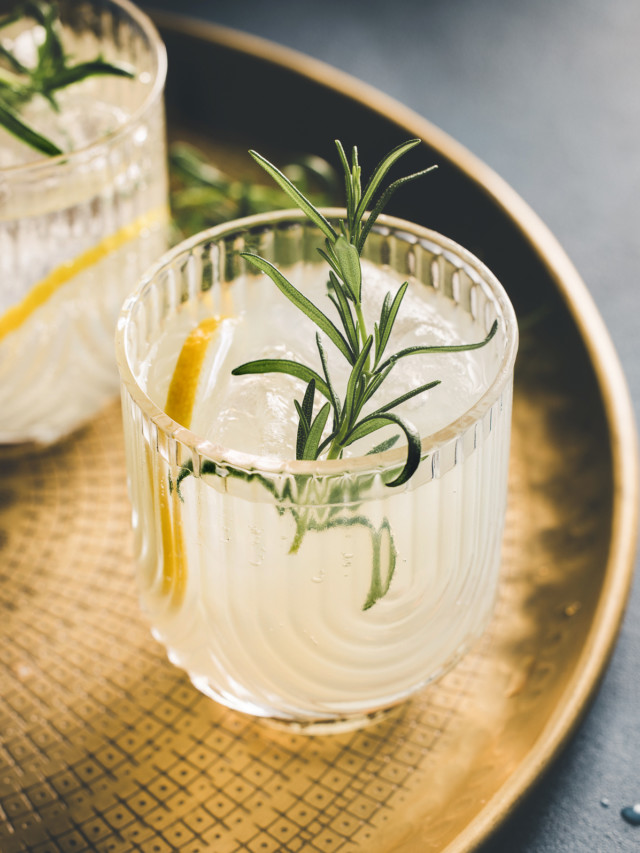 two glasses of a gin sour cocktail on a gold tray garnished with rosemary and lemon
