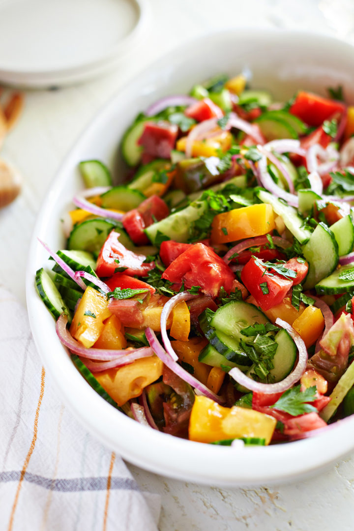 salad with onions, tomatoes, and cucumbers in a white dish on a table next to plates