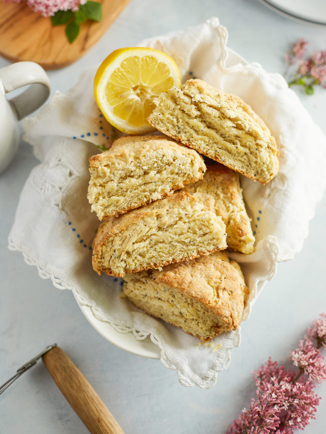 A basket lined with a tea towel filled with golden, flaky lemon poppy seed scones, surrounded by fresh-cut flowers. These tender scones are made with buttermilk for extra softness.