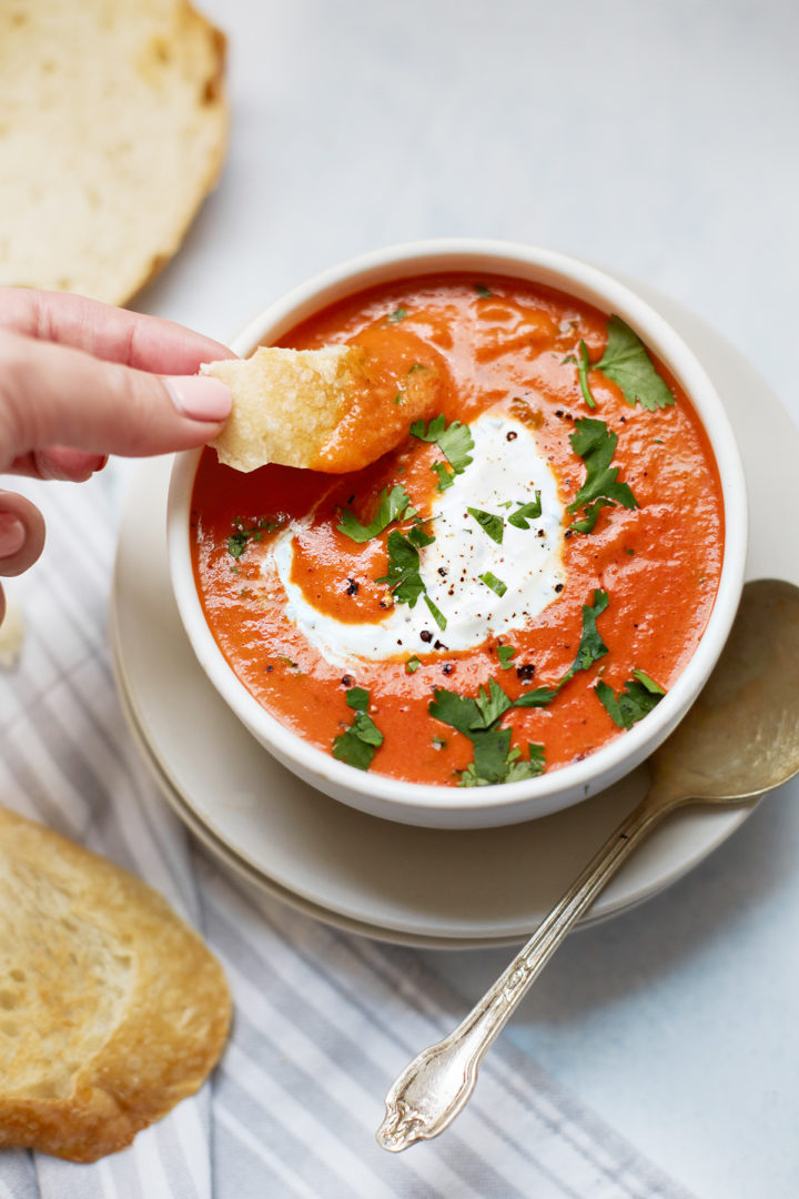 woman dipping bread in a bowl of roasted red pepper soup swirled with cilantro cream