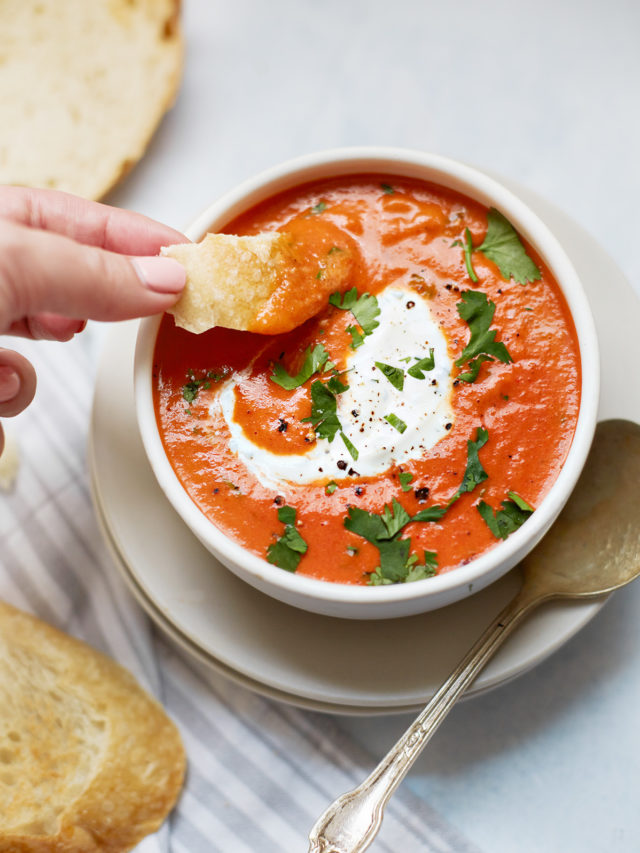 woman dipping bread in a bowl of roasted red pepper soup swirled with cilantro cream