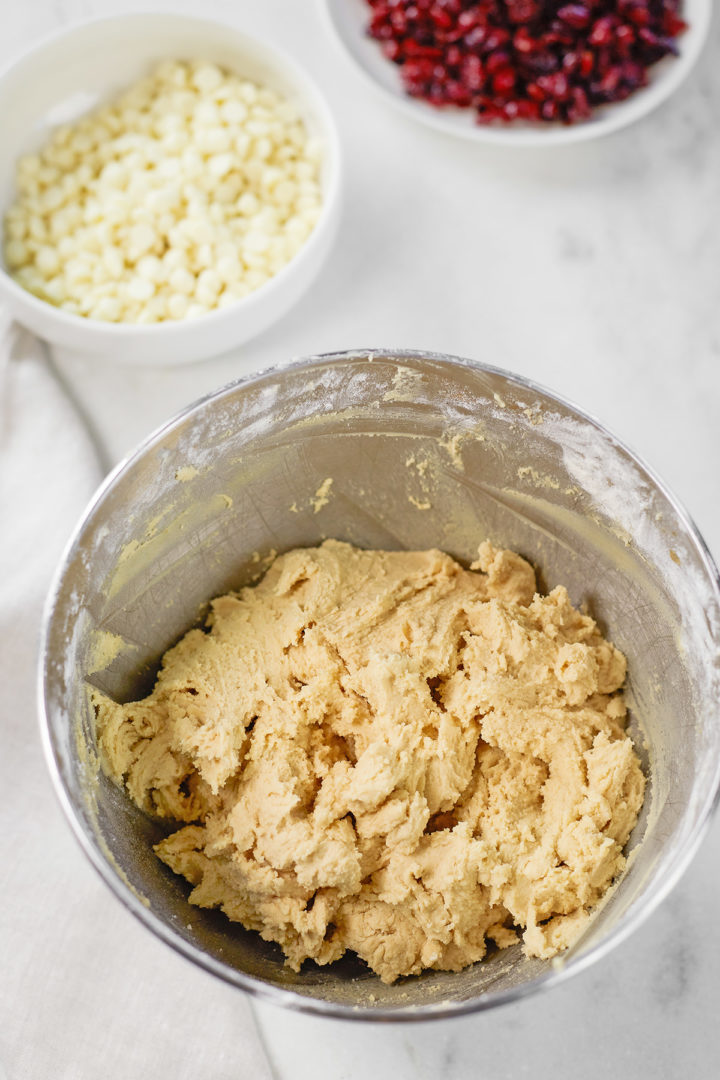 a bowl of cookie dough next to bowls of white chocolate chips and dried cranberries