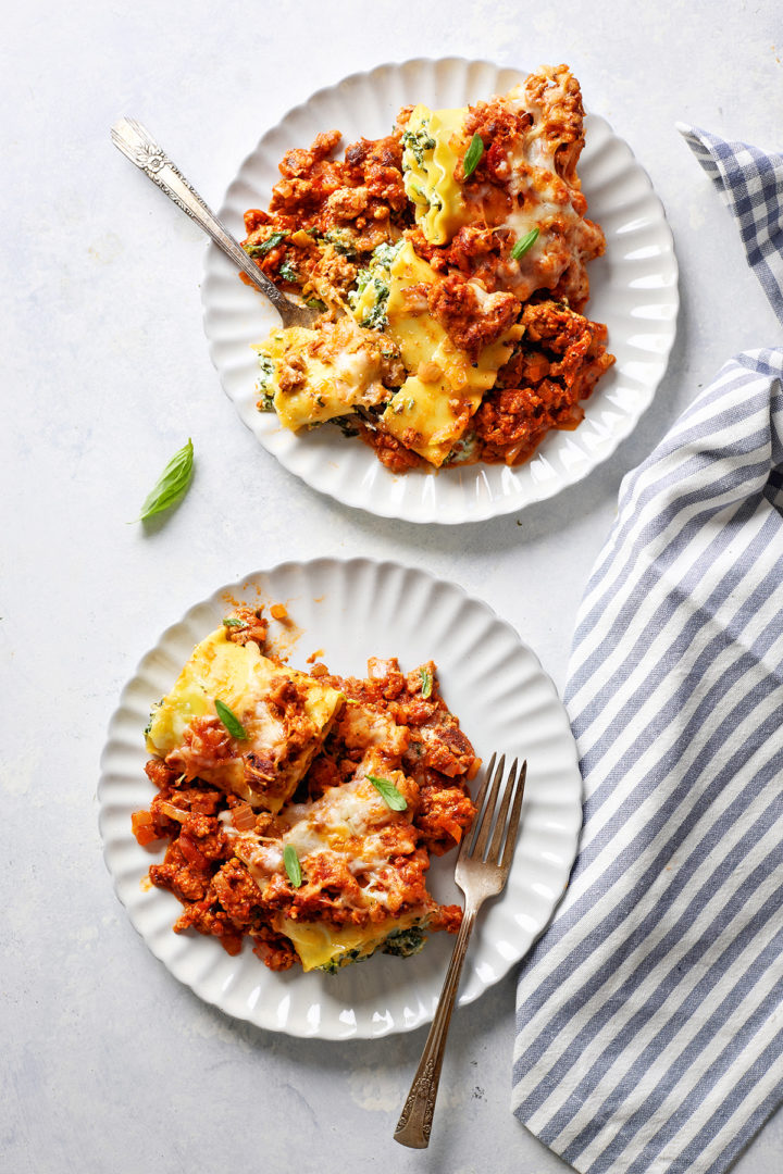 Two plates of sausage lasagna rolls served on white plates with forks, garnished with fresh basil. A bite has been taken from one piece, revealing the cheesy spinach filling.