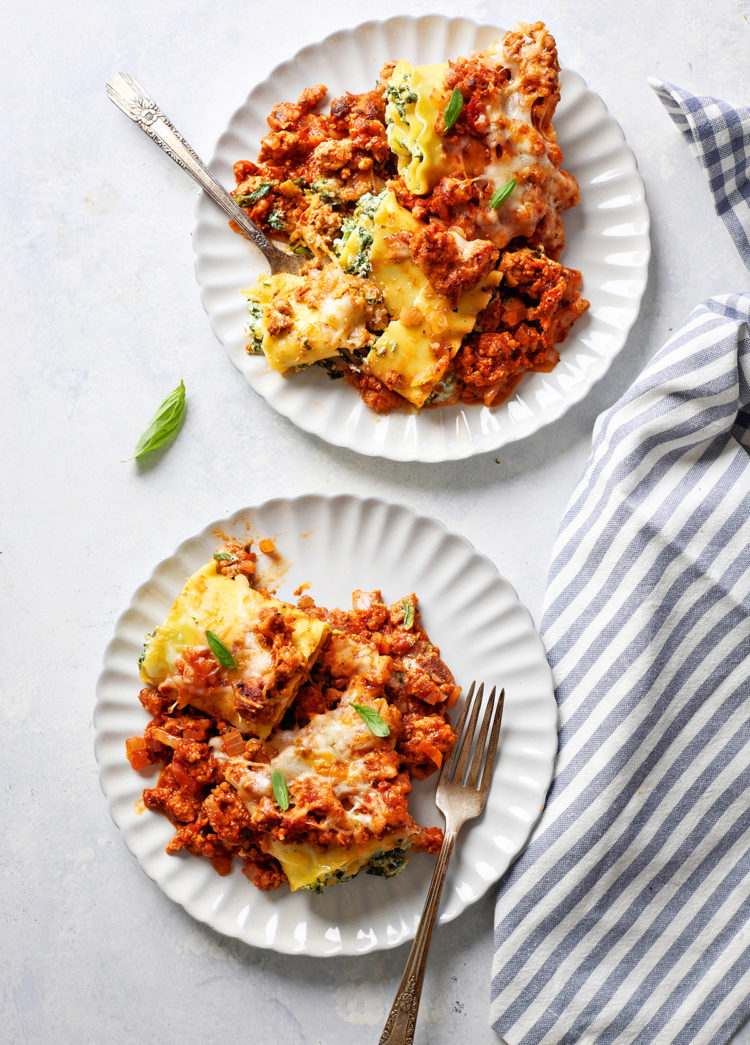 Two plates of sausage lasagna rolls served on white plates with forks, garnished with fresh basil. A bite has been taken from one piece, revealing the cheesy spinach filling.